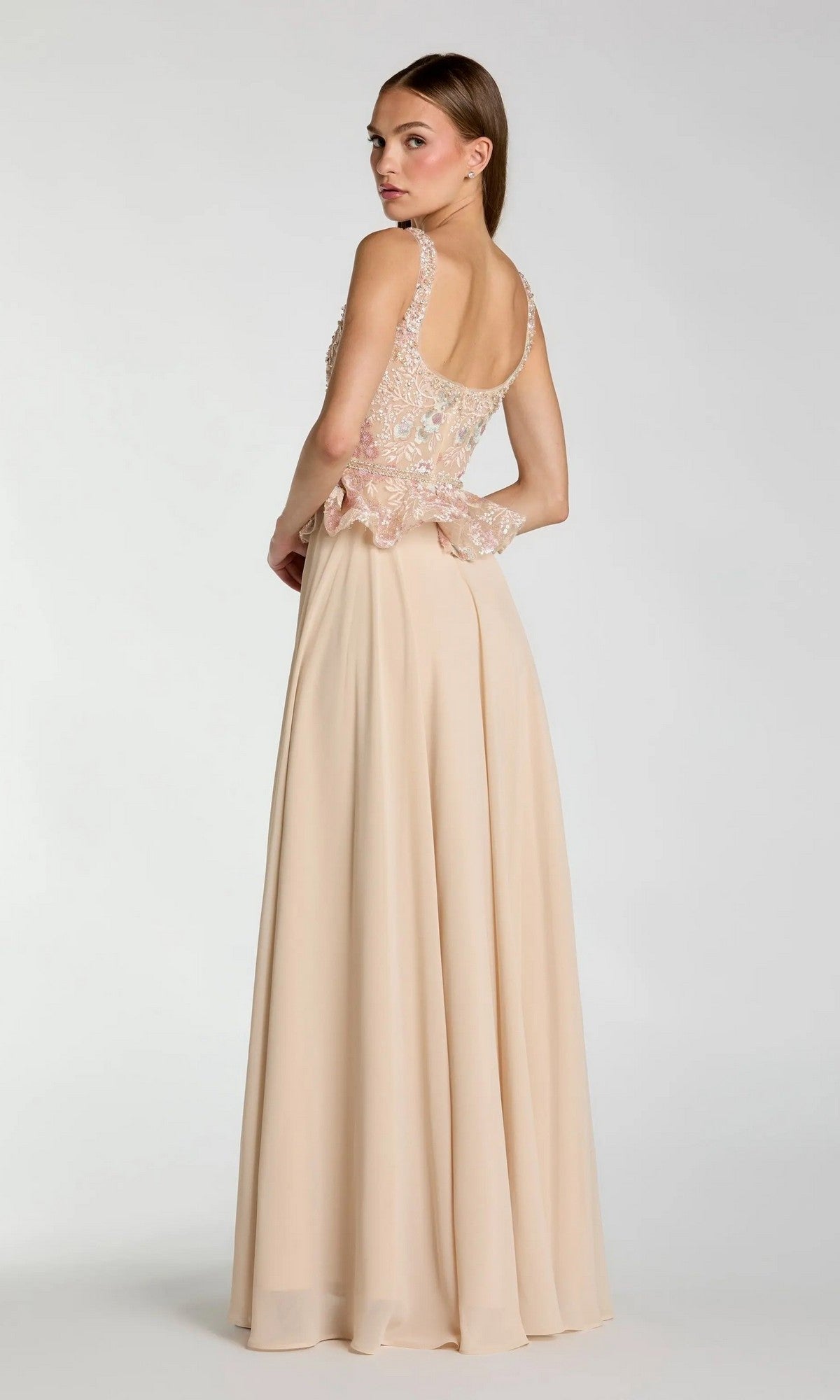 A woman wearing the Mac Duggal 21317 long formal dressโan elegant, floor-length, sleeveless beige A-line chiffon evening gown with a floral embroidered bodice and sophisticated peplum waistlineโstands against a plain light background.